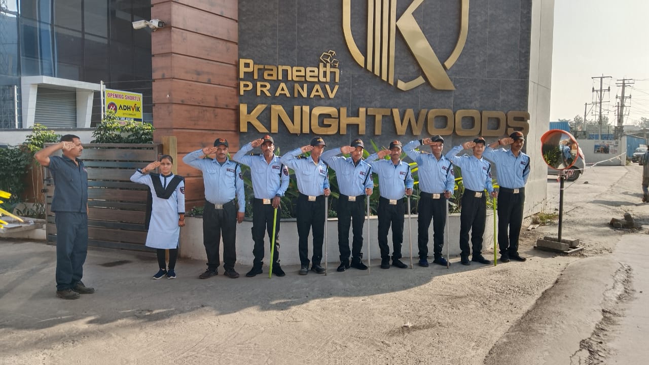 "A group of nine security personnel and one child stand in a line, saluting in front of a building with a sign that reads 'Praneeth Pranav Knightwoods.' The security staff are dressed in light blue shirts, dark pants, and caps, while the child wears a white and black uniform. The background features a modern building with large windows, a sign for 'ADHViK' indicating an upcoming opening, and a convex mirror on the right side."