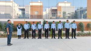 "Nine individuals, predominantly dressed in light blue shirts and black pants, stand in a straight line outdoors, holding batons. One person in a white outfit stands among them. Another individual in a darker blue uniform stands apart, facing the group. The setting features a modern building with reflective glass, a brown and white facade, and nearby greenery."