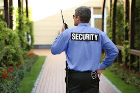 A security guard is walking down a pathway lined with greenery. The guard is wearing a blue uniform with the word "SECURITY" printed in large white letters on the back. The guard is holding a walkie-talkie to the face and has a key ring attached to the belt. The face of the guard is pixelated for privacy.