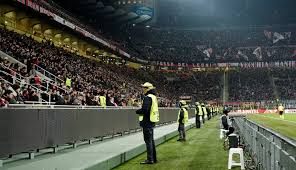This image captures a crowded stadium during what seems to be a significant sporting event, likely a football (soccer) match. The presence of security personnel and the packed stands highlight the scale of the event and the measures taken to ensure safety and order.