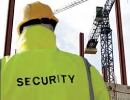 A person wearing a yellow high-visibility jacket with the word "SECURITY" written on the back and a yellow hard hat is standing at a construction site. In the background, there is a large construction crane and some steel beams.