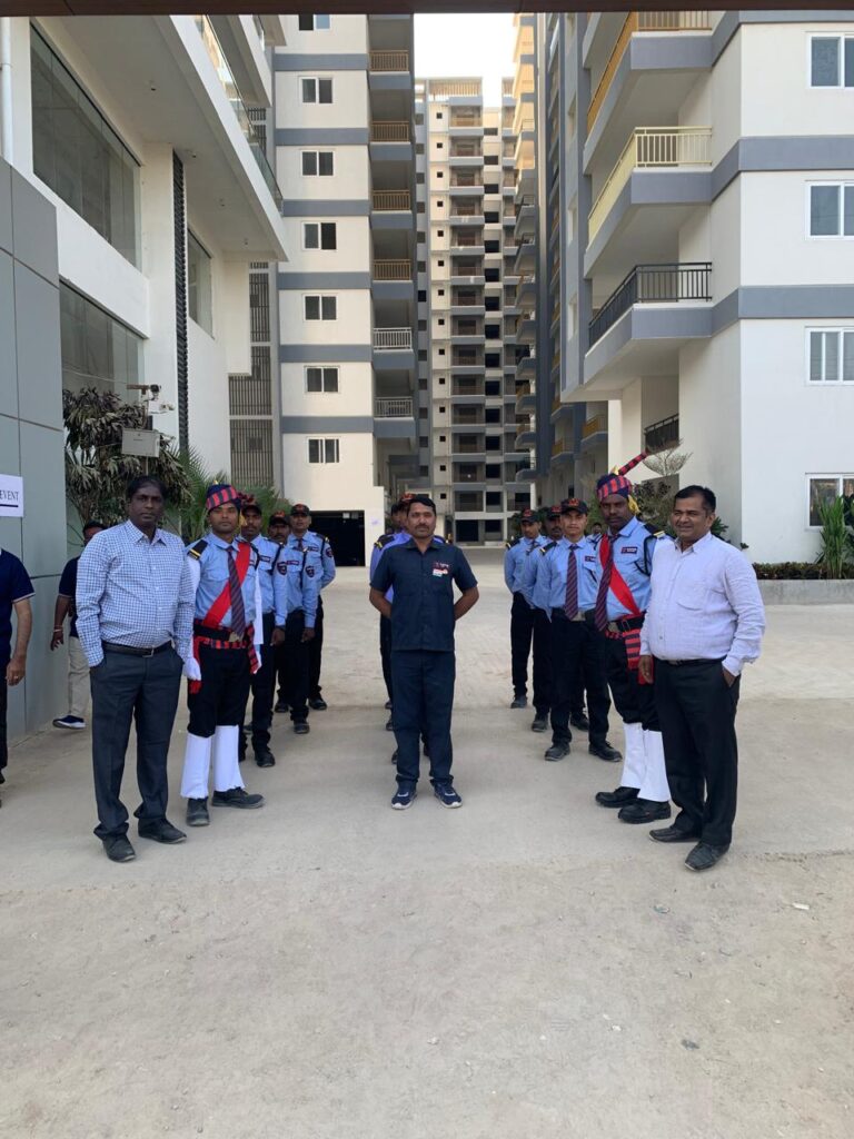 A group of people stands in a semi-circle formation between two tall residential buildings in an outdoor area. The group includes individuals in uniforms, possibly security personnel, and others in casual or business attire. The background shows multiple floors of buildings with balconies and windows. The scene appears to be a formal gathering or inspection.