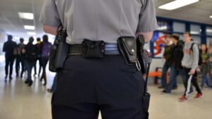 This image shows a security officer monitoring students as they walk through a school hallway. It highlights the presence of security personnel in educational institutions, emphasizing school safety and security measures.